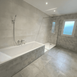 Salle de bain rénovée avec carrelage gris grand format, baignoire et douche à l’italienne.