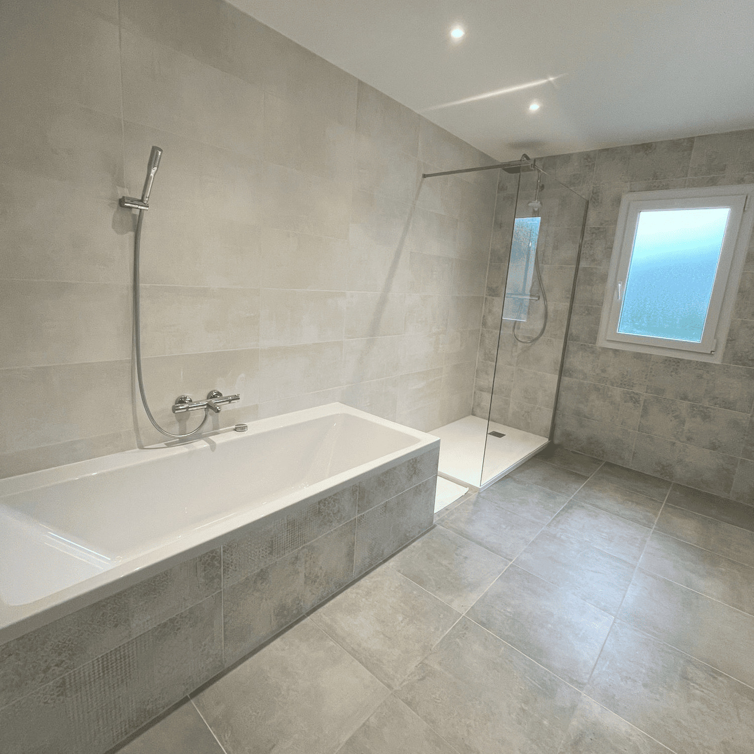 Salle de bain rénovée avec carrelage gris grand format, baignoire et douche à l’italienne.