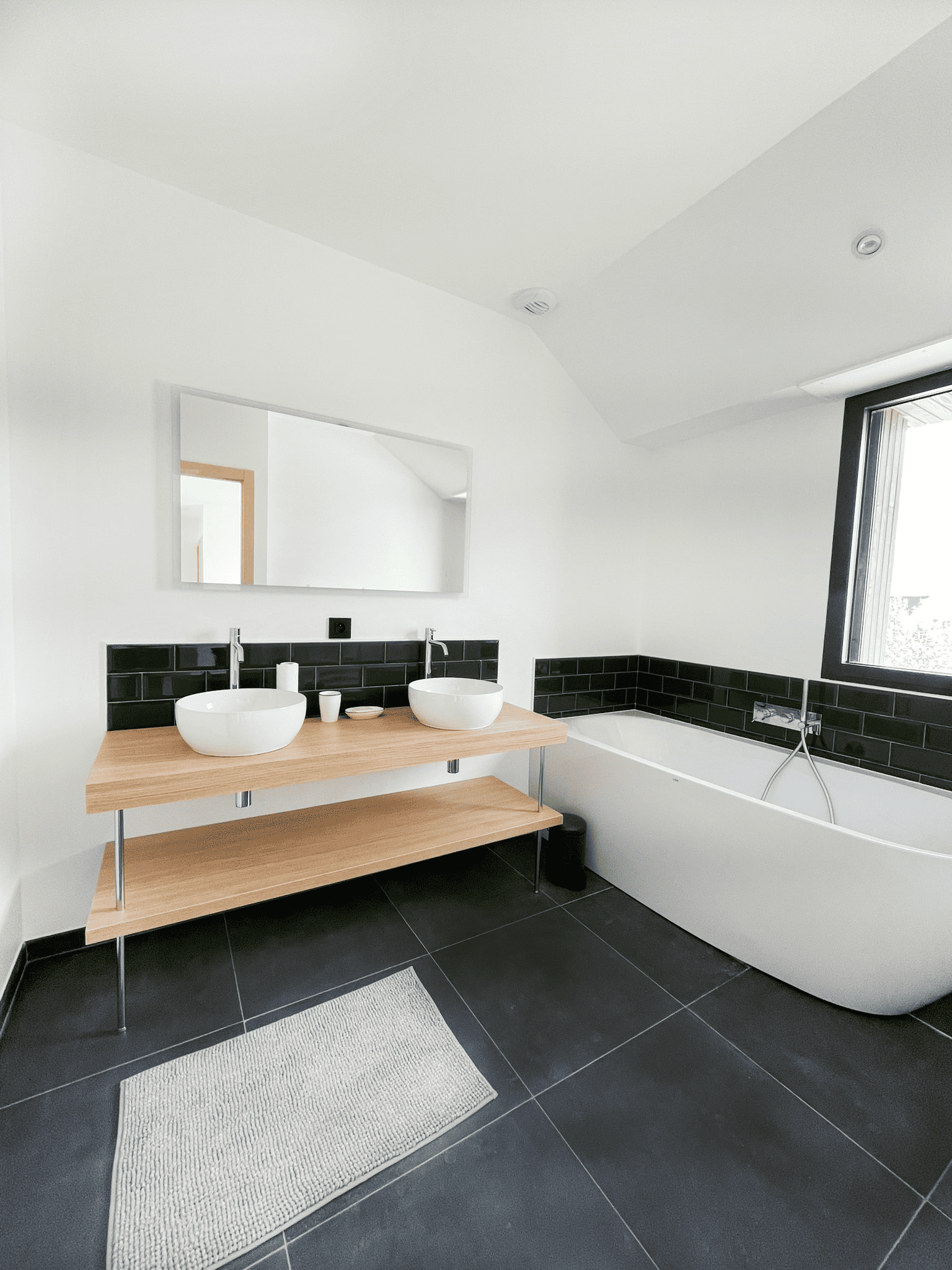 Salle de bain contemporaine rénovée avec carrelage noir, baignoire îlot et vasques posées sur plan de travail en bois massif